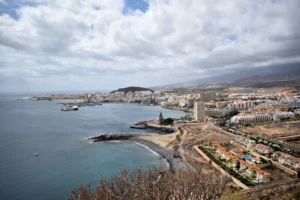 High-angle landscape view from the Coastal Montaña Guaza Outlook showing the Los Cristianos coastline, the port with a docked ferry, and Playa de las Vistas under a cloudy sky in Tenerife.